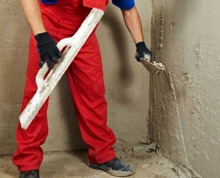 MDS-bautenschutz-bausanierung-frankfurt-kellerwaende-trockenlegen Handwerker in roter Arbeitskleidung verputzt feuchte Kellerwand – Sanierungsarbeit zur Trockenlegung und Abdichtung von Innenwänden.